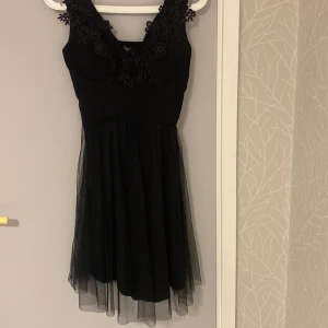 Svart kort klänning med spetsdetaljer - Supersnygg svart kort klänning från Sisters Point med v-ringning och blommiga spetsdetaljer upptill. Kjolen är i flera lager tunn tyll som ger en luftig känsla. Perfekt för dig som vill sticka ut med en elegant och ungdomlig look.