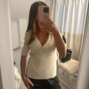 Ribbad omlottopp i cremevitt - Säljer en ribbad omlottopp i cremevitt med v-ringning och korta ärmar. Toppen har en figurnära passform och omlottdesign som ger en snygg siluett. Perfekt att styla med både jeans och kjol för en trendig look.