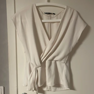 Vit omlottopp från Zara - Säljer en stilren vit omlottopp från Zara med v-ringning och knyt i midjan. Toppen har korta ärmar och är tillverkad i ett mjukt, lätt material som ger ett snyggt fall. Perfekt för dig som gillar en clean och fräsch look.💞