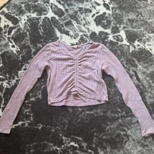 Ljuslila croppad ribbad topp - Säljer en ljuslila croppad topp med långa ärmar och v-ringning. Toppen har snygg snörning framtill och är ribbad med ett diskret randigt mönster. Materialet är mjukt och stretchigt, perfekt för en chill och trendig look.