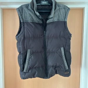 Svart dunväst från BOSS med grå detaljer - Snygg svart dunväst från BOSS med grå axelparti och krage. Västen har dragkedja framtill och två fickor med grå kant. Perfekt för lager-på-lager och stilren look. Klassisk passform och quiltad design.