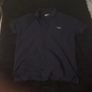 Mörkblå pikétröja från Lacoste - Klassisk mörkblå pikétröja från Lacoste med korta ärmar och ikoniska krokodilloggan på bröstet. Tröjan har krage och knäppning framtill, tillverkad i mjuk bomull som känns skön mot huden. Perfekt för en clean och sportig stil.