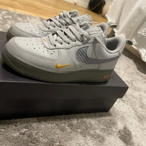 Nike Air Force 1 grå med orange detaljer - Säljer ett par grå Nike Air Force 1 med snygga orange detaljer på swoosh och AIR-loggan. Skorna har klassisk låg siluett, snörning och är tillverkade i skinn och textil. Perfekta för dig som gillar stilrena sneakers med en twist.
