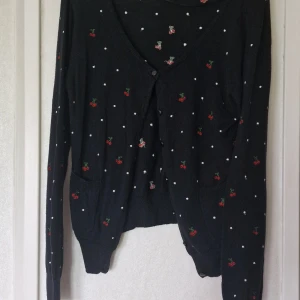 Svart kofta med körsbär från R&D - Svart kofta från R&D med broderade körsbär och små vita prickar över hela plagget. V-ringad med knappar framtill, två fickor och långa ärmar. Tunn och stickad, perfekt för lager-på-lager. Vid höger ficka kan man se att en maska är skadad men några andra skavanker har jag inte sett och det är inget som stör helhetsintrycket.