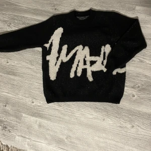 Svart stickad tröja med vit text - Säljer en svart stickad tröja med stor vit text i graffiti-stil över hela framsidan. Tröjan har rund halsringning och långa ärmar. Perfekt för dig som gillar streetwear och vill sticka ut med en cool design.