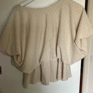 Beige blus med volang och v-ringning - Säljer en luftig beige blus med v-ringning och korta, vida ärmar. Blusen har en subtil struktur i tyget och en volang nedtill som ger en avslappnad och trendig look. Perfekt att styla med jeans eller kjol för en fräsch outfit.