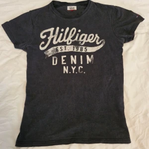 Mörkblå t-shirt Tommy Hilfiger Denim - Snygg mörkblå t-shirt från Tommy Hilfiger Denim med stort vitt tryck på bröstet och klassisk rund hals. Kortärmad modell i mjuk bomull, perfekt för en chill streetstyle-look. Liten Hilfiger-logga broderad på ärmen för extra detalj.
