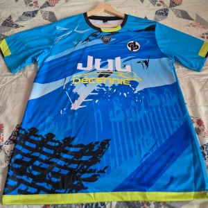 JUL 90 minutes jersey  - Snygg blå JUL jersey med coolt grafiskt tryck i svart, vitt och gult. Kortärmad modell med svarta och gula detaljer vid krage och ärmslut. Materialet är lätt och andas, perfekt för match eller träning. Tryck på både fram- och baksida med text och loggor.