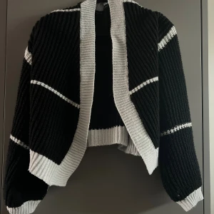 Svartvit stickad kofta med ribbade detaljer - Säljer en svart stickad kofta med breda vita ribbade kanter och vita ränder på ärmar och kropp. Koftan är något kortare och har en öppen framsida utan knappar. Den har ganska tjockt, mysigt material och är i storlek S