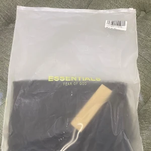 Svart Essentials Fear of God t-shirt - Den är kopia1:1 Svart t-shirt från Essentials Fear of God i bomull med klassisk passform och korta ärmar. T-shirten har tryckt logga på bröstet och rund halsringning. Perfekt för en clean och stilren streetwear-look. 