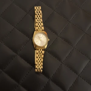 Guldig klocka från Regal med länkarmband - Stilren klocka från Regal i guldig färg med klassiskt länkarmband och rund urtavla. Urtavlan är ljus med tydliga markeringar och guldfärgade visare. Klockan har en räfflad kant runt urtavlan och smidig låsmekanism på armbandet. Använt få gånger, tagit av länkar som jag tyvärr har slängt men det går säkert att köpa några nya! Drar ner priset eftersom inte alla länkar är med. Köparen står för frakt :)