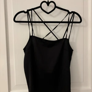 Svart satin topp från  H&M Divided - Svart linne från H&M Divided i satinliknande material. Linnet har tunna axelband som korsas i ryggen för en snygg detalj. Enkel och stilren modell som sitter snyggt på kroppen.