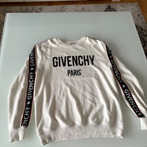 Vit Givenchy sweatshirt med logga - Snygg vit sweatshirt från Givenchy med svart logga framtill och svarta band med Givenchy-tryck längs ärmarna. Tröjan har rund halsringning, ribbade muddar och är tillverkad i mjuk bomull. Perfekt för dig som vill ha en clean och trendig look.