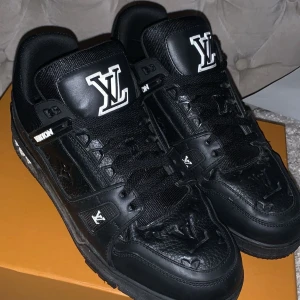 Louis Vuitton svarta sneakers i skinn - Svarta Louis Vuitton sneakers i skinn med tydliga LV-loggor på plös, häl och tå. Skorna har snörning, platt sula och detaljer i vitt. Tillverkade i Italien och har en chunky, mer som sportig vibe med exklusiv känsla. Dem kommer med kartong, kvitto och nästan alla tillbehör, plus får man en extra vit-snöre 