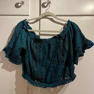 Mörkgrön offshoulder topp H&M Divided - Säljer en mörkgrön croppad offshoulder topp från H&M Divided. Toppen har korta volangärmar, elastisk kant upptill och nertill samt dekorativa spetsdetaljer framtill och på ärmarna. Perfekt för dig som gillar boho-vibbar och snygga detaljer.