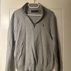 Grå stickad half zip från Polo Ralph Lauren - Grå stickad tröja från Polo Ralph Lauren med half zip och klassisk broderad logga på bröstet. Tröjan har lång ärm, ribbade muddar och hög krage. Perfekt för lager-på-lager och snygg till jeans eller chinos.