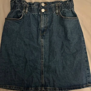 Jeanskjol med resårmidja från Lindex - Snygg blå jeanskjol från Lindex med resår i midjan och dubbla knappar framtill. Kjolen har klassiska fickor både fram och bak samt bälteshällor. Perfekt för dig som gillar en avslappnad och trendig stil.