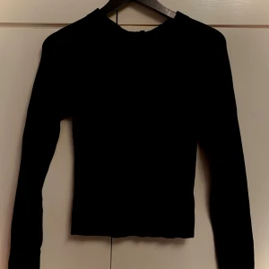 Svart ribbad långärmad topp med knyt bakom - Svart ribbad långärmad topp med öppen rygg och knytband i nacken. Snygg och figurnära modell. Materialet är stretchigt och mjukt, vilket gör att den passar S/M/L. 