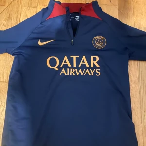 PSG Nike blå fotbollströja L - Säljer en snygg Paris Saint-Germain fotbollströja från Nike i marinblått med röda detaljer och guldiga loggor. Tröjan har kort dragkedja vid halsen, Qatar Airways-tryck och är tillverkad i Dri-FIT-material som andas. Perfekt för dig som älskar fotboll och PSG.