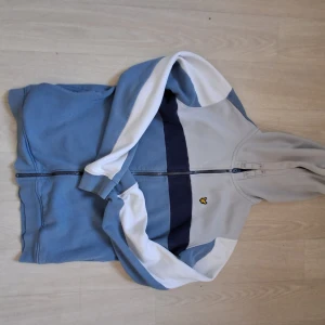 Blå och grå hoodie zipper från Lyle & Scott - Snygg hoodie från Lyle & Scott i blått, grått och vitt med dragkedja framtill och klassisk gul logga på bröstet. Tröjan har huva, långa ärmar och är tillverkad i mjuk bomullsmix. Perfekt för chill dagar eller när du vill ha en sportig look.