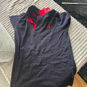 Mörkblå pikétröja från Ralph Lauren - Snygg svart pikétröja från Ralph Lauren med röd krage och broderad logga på bröstet. 