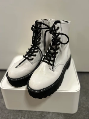 Vita boots med platåsula och snörning - Säljer ett par vita boots med svart snörning och grov platåsula. Skorna har rund tå, dragkedja på sidan och kontrastsydda detaljer. Materialet är skinnimitation och sulan är rejält mönstrad för extra edge.