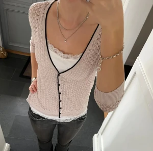 Ljusrosa stickad blus med svarta knappar - Supersöt ljusrosa stickad blus med v-ringning och svarta knappar framtill. Blusen har trekvartsärm och svart kant längs urringningen. Perfekt att styla med ett linne under för en chic look.