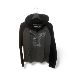 Grå och svart hoodie American Eagle - Säljer en grå och svart hoodie från American Eagle med stor örnprint på bröstet. Tröjan har huva med snörning, känguruficka och långa svarta ärmar. Mjuk fleece på insidan och klassisk passform. Perfekt för chill dagar och streetstyle.