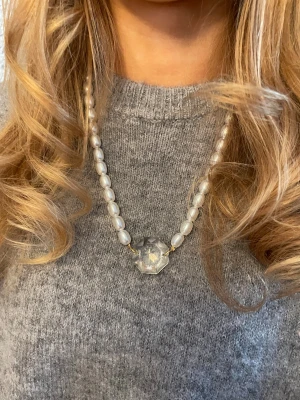 Pärlhalsband med stor kristall - Unikt halsband med vita ovala pärlor och en stor, multifasetterad kristall i mitten. Pärlorna har en fin lyster och kristallen ger ett coolt blänk. Guldfärgade detaljer vid kristallen. Perfekt statement-piece för dig som gillar att sticka ut.