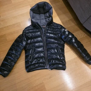 Svart pufferjacka från Colmar - Snygg svart pufferjacka från Colmar med glansig finish och vadderad design. Jackan har huva, dragkedja framtill och två fickor med dragkedja. Perfekt för kyliga dagar och ger en modern look. Passar dig som gillar en stilren och enkel look..