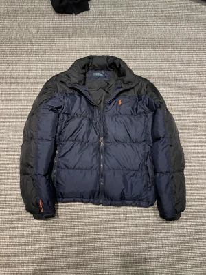 Mörkblå pufferjacka från Polo Ralph Lauren - Snygg mörkblå pufferjacka från Polo Ralph Lauren med orange logga på bröstet. Jackan har hög krage, dragkedja framtill och två sidofickor med dragkedja. Materialet är syntet och jackan har en loose passform, perfekt för kalla dagar. Ens dragkedjan är sönder 