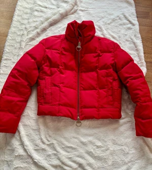 Röd pufferjacka från SAND - Säljer en fet röd pufferjacka från SAND med coola ringformade dragkedjor och hög krage. Jackan är quiltad och har två fickor framtill. jackan är croppad i modellen, perfekt för kalla dagar.
