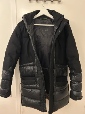 Svart pufferjacka från BOSS - Säljer en svart vinterjacka från BOSS med huva och coolt quiltad design. Jackan har både matta och blanka partier, flera fickor framtill och dragkedja med knappslå. Perfekt för kalla dagar och har en modern, urban vibe.