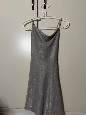Glittrig silver klänning med öppen rygg - Supersnygg glittrig silverfärgad kortklänning med tunna axelband och draperad urringning. Klänningen har öppen rygg med knytband och är figurnära upptill med lätt utställd kjol. Perfekt för dig som vill sticka ut och glänsa. Aldrig använd utan endast testad då jag är mer guldperson😅 superfin till nyår eller annat festligt tillfälle! 🩷