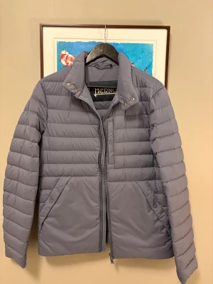 Grå dunjacka från Herno - Snygg grå dunjacka från Herno med quiltad design och hög krage. Jackan har dragkedja framtill, tryckknappar vid kragen och två stora fickor. Perfekt för kyliga dagar och har en stilren look som funkar till det mesta. Använd en gång och nypris flera tusen.