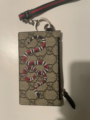 Gucci plånbok med orm och dragkedja - Snygg plånbok från Gucci med klassiskt GG-mönster i beige och brunt, dekorerad med en röd, vit och svart orm på framsidan. Plånboken har en dragkedja och en handledsrem i grönt och rött. Insidan är svart och har präglad Gucci-logga.