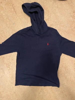 Marinblå hoodie från Polo Ralph Lauren - Snygg marinblå hoodie från Polo Ralph Lauren med klassisk röd broderad logga på bröstet. Tröjan har huva och långa ärmar, tillverkad i mjuk bomull som är skön mot huden. Perfekt för dig som gillar stilrena och tidlösa plagg.
