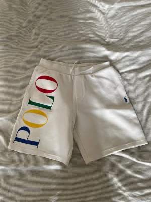 Vita Polo Ralph Lauren shorts - Säljer ett par vita shorts från Polo Ralph Lauren med stor färgglad POLO-logga längs ena benet och blå broderad logga på andra sidan. Mjukt material och snörning i midjan för skön passform. Perfekta för sommardagar och chill vibes.