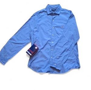 Blå skjorta från Polo Ralph Lauren - Klassisk blå skjorta från Polo Ralph Lauren med lång ärm och knappar framtill. Skjortan har en broderad logga på bröstet och är tillverkad i mjuk bomull. Perfekt för en stilren och avslappnad look.