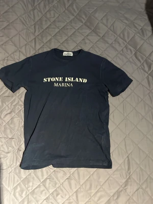 Stone Island Marina t-shirt marinblå - Marinblå t-shirt från Stone Island Marina med vit logga tryckt på bröstet. Klassisk rund halsringning och korta ärmar. Tillverkad i mjuk bomull för en skön känsla. Perfekt för dig som gillar streetwear och vill ha en clean look.