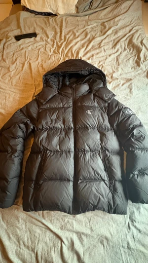 Svart pufferjacka från Calvin Klein Jeans - Svart pufferjacka från Calvin Klein Jeans med huva och dragkedja framtill. Jackan har diskret CK-logga på bröstet och ärmen, två fickor med dragkedja och quiltad design. Perfekt för kalla dagar och riktigt snygg till streetwear-stil.
