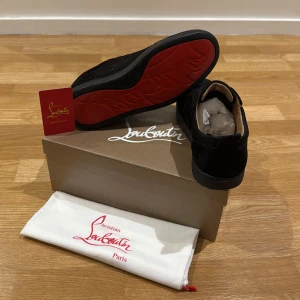 Christian Louboutin svarta sneakers mocka - Snygga svarta sneakers från Christian Louboutin i lyxig mocka med ikonisk röd sula. Skorna har rund tå, platt sula och klassisk snörning. Perfekta för dig som vill sticka ut med exklusiv stil och detaljer. Kommer med originalpåse och box.