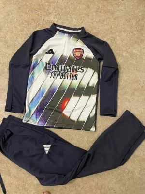 Arsenal träningsset från Adidas - Säljer ett Arsenal träningsset från Adidas med långärmad tröja och matchande byxor. Tröjan har klubbmärke, Adidas-logga och Emirates Fly Better-tryck, samt ett coolt randigt mönster i vitt, svart och färgdetaljer. Byxorna är svarta med Adidas-logga. Perfekt för fotbollsträning.