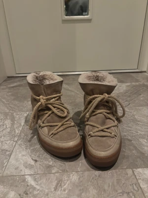 Beige moon boots från Inuikii - Säljer ett par beige moon boots från Inuikii med fluffigt foder och grova snören. Skorna har en rund tå, platt sula och är tillverkade i mocka och skinn. Perfekta för kalla vinterdagar med sin varma insida och snygga design.