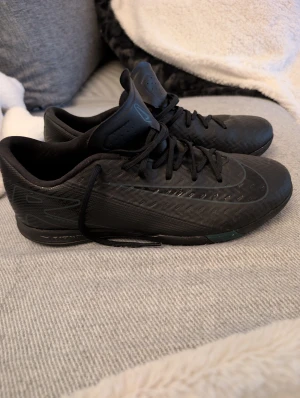 Svarta Nike fotbollsskor strl 42 - Säljer ett par svarta Nike fotbollsskor med snyggt mönstrad ovandel och diskret Nike-logga på sidan och sulan. Skorna har snörning och en låg profil, perfekt för snabba rörelser på planen. Materialet är syntet och designen är riktigt sportig och modern. Använda inomhus fint skick