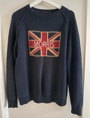 Mörkblå stickad tröja Morris M - Snygg mörkblå stickad tröja från Morris i storlek M. Framsidan har ett broderat Union Jack-motiv med Morris-loggan i rött, beige och blått. Tröjan har ribbade muddar vid hals, ärmslut och nederkant samt långärmad passform. Perfekt för dig som gillar klassisk stil med en twist.