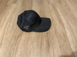 Svart keps från Prada i nylon - Svart keps från Prada med klassisk böjd skärm och ikonisk metalltriangel med logga på sidan. Tillverkad i slät nylon som ger en clean och modern look. Perfekt för dig som vill ha en stilren accessoar med lyxig känsla.