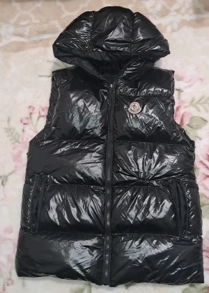 Svart dunväst från Moncler med huva - Snygg svart dunväst från Moncler med glansig finish och huva. Västen har dragkedja framtill, två fickor med dragkedja och Moncler-logga på bröstet. Perfekt för lager-på-lager och en cool streetstyle-look.