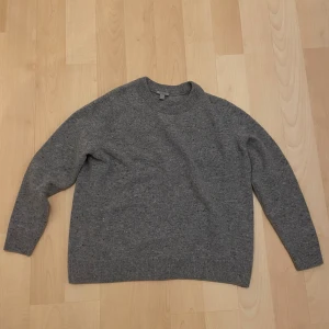 Grå stickad tröja från COS - Mysig grå stickad tröja med rund hals. Tröjan har en enkel design med ribbade muddar vid hals, ärmslut och nederkant. Perfekt för kyliga dagar och passar dig som gillar en klassisk look. Materialet känns mjukt och värmande. i mycket gott skick, inga defekter