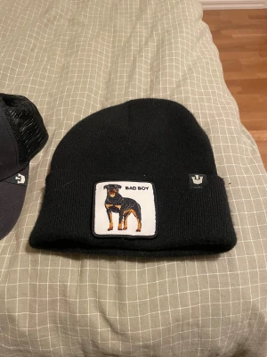 Svart mössa Bad Boy från Goorin Bros - Svart ribbstickad mössa från Goorin Bros med patch framtill som visar en rottweiler och texten 'BAD BOY'. Klassisk form med uppvikt kant och liten logga på sidan. Perfekt accessoar för en edgy streetstil.
